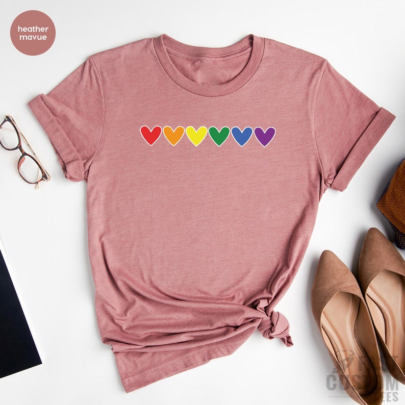 May include: A heather mauve t-shirt with a row of rainbow hearts across the chest. The hearts are outlined in white.