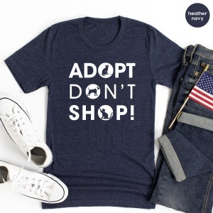 May include: A heather navy blue t-shirt with the text "ADOPT DON'T SHOP!" in white. The text is stacked vertically with a black cat, dog, and cat silhouette in circles between the words.