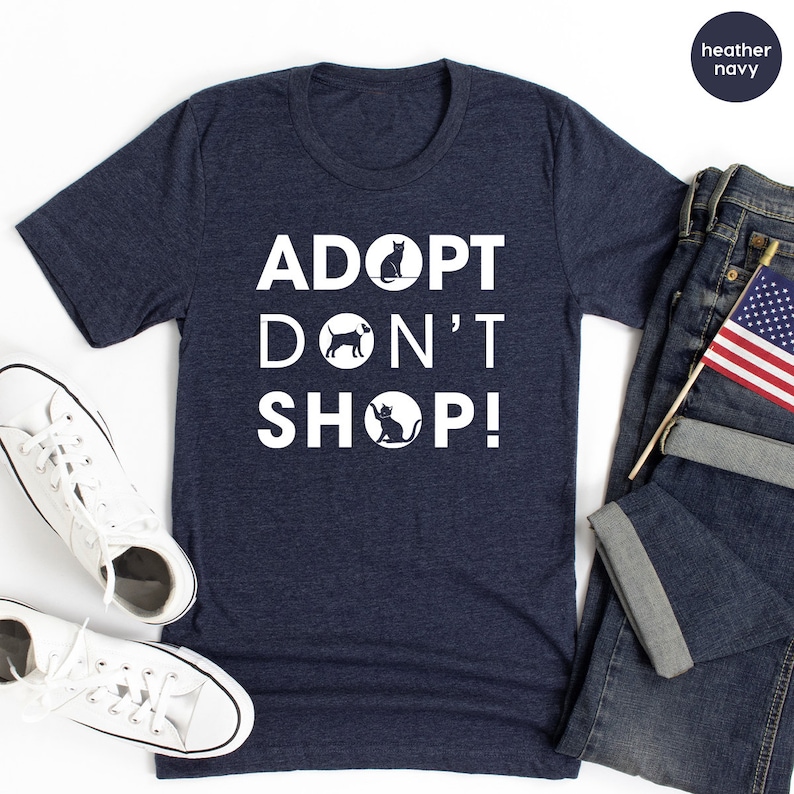 May include: A heather navy blue t-shirt with the text "ADOPT DON'T SHOP!" in white. The text is stacked vertically with a black cat, dog, and cat silhouette in circles between the words.