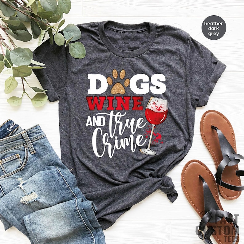 May include: Heather dark grey t-shirt with the text "Dogs, Wine, and True Crime" in white and red. The design includes a paw print, a wine glass with red wine, and the text "Fast Custom Tees". The shirt is paired with blue jeans and sandals.