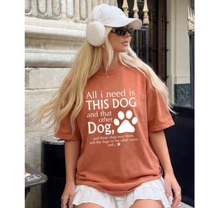 A woman wearing a white cap and sunglasses, with a t-shirt that says "All I need is this dog and that dog over there. And those dogs over there and the dogs in the other room."