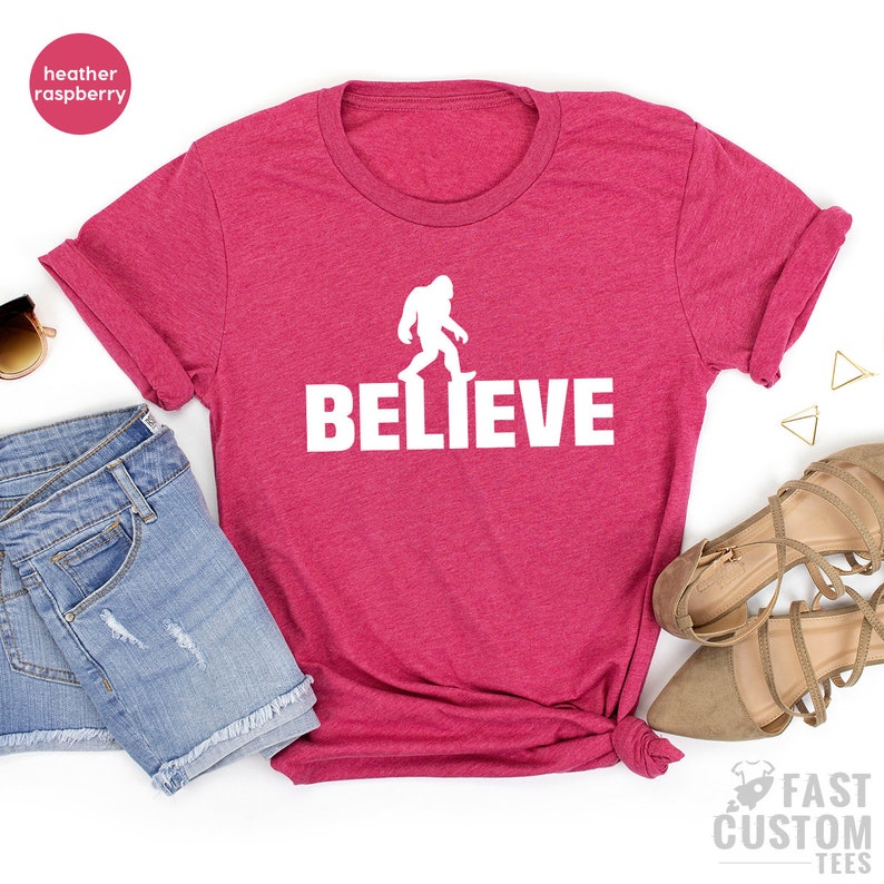 May include: A heather raspberry t-shirt with a white silhouette of Bigfoot walking and the word "BELIEVE" printed below it.