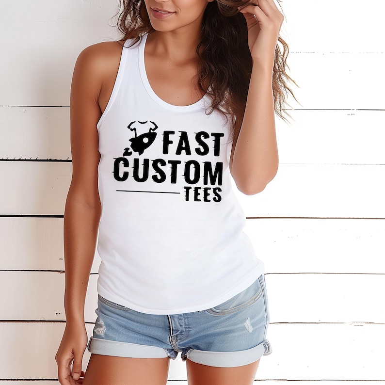 May include: White racerback tank top with the text "FAST CUSTOM TEES" in black. The graphic includes a rocket ship and a baby onesie. The tank top is paired with denim shorts.