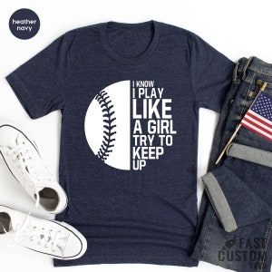 Op de afbeelding: Een heideblauwe t-shirt met een witte baseball-afbeelding en de tekst "I know I play like a girl try to keep up".
