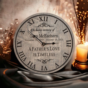 May include: A gray wooden clock with Roman numerals and black hands. The clock face reads "In loving memory of Gary McEachern November 21, 1957 - October 28, 2023 A Father's Love Is Timeless".