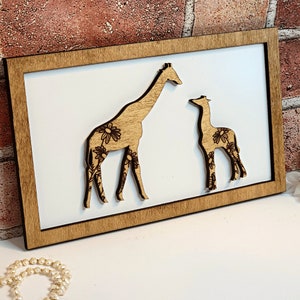 May include: A wooden wall decor featuring a cutout of two giraffes with floral patterns. The giraffes are facing each other, with the larger giraffe on the left and the smaller giraffe on the right. The decor is mounted on a brown wooden frame.