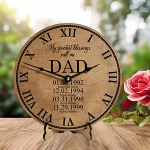 May include: A wooden clock with Roman numerals and a stand. The clock face has the words "My greatest blessings call me DAD" and a list of names and dates.
