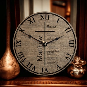 May include: A wooden clock with Roman numerals and a cross design. The clock face has the name "Matthew" and a list of bible verses. The clock face reads "You are... chosen (1 Peter 2:9) made for a purpose (Ephesians 2:10)  workmanship of God (John 1:12) his workmanship (Ephesians 2:10) equipped with strength for the battle (Psalm 18:39) heated by His stripes (Isaiah 53:5) forgiven and set free (Ephesians 1:7)"