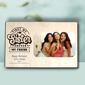 May include: A wooden photo frame with a brown background and a white border. The frame has a cutout for a photo and a black and white graphic that says "First My Sister Forever My Friend" with hearts and stars. The text "Happy Birthday! Love, Jenna 2023" is also included.