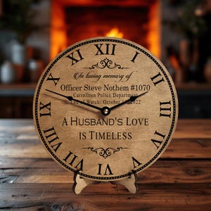 May include: A wooden clock with Roman numerals and a black stand. The clock face reads "In loving memory of Officer Steve Nothem #1070 Carrollton Police Department End of Watch: October 19, 2022 A HUSBAND'S LOVE IS TIMELESS".