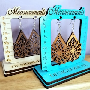 May include: Two wooden earring display stands with a ruler marked in half inches up to 4 1/2 inches. The stands are painted in turquoise and white. The earrings are brown and feature a mandala design. The stands are labeled "DESIGNODEAL" and "Measurements".