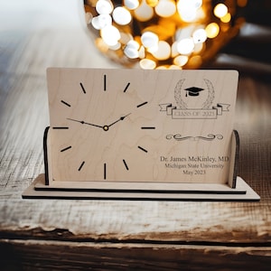 May include: A wooden desk clock with a black clock face and hands. The clock has a graduation cap and laurel wreath design with the text "Class of 2023". The clock also has the text "Dr. James McKinley, MD, Michigan State University, May 2023".