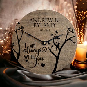 May include: A wooden clock with a black silhouette of two trees and a bird. The clock face reads "I am always with you" with two small hearts. The clock is personalized with the name "Andrew R. Ryland" and the dates "1947 - 2023".