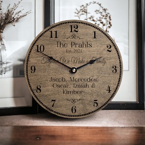 May include: A round wooden clock with black hands and a brown wood grain finish. The clock face has black numbers and text that reads "The Prahls Est. 2021 Together We Make a Family Jacob, Mercedes, Oscar, Izaiah & Kimber".