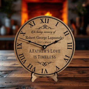May include: A wooden clock with Roman numerals and a black clock face. The clock face reads "In loving memory of Robert George Lapsansky July 18, 1941 - March 17, 2021 A Father's Love Is Timeless".