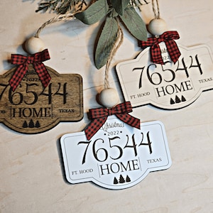 May include: Three wooden Christmas ornaments with red and black plaid bows. Each ornament features a house-shaped sign with the text "76544 HOME" and the location "FT. HOOD TEXAS". One ornament is brown wood, and the other two are white.