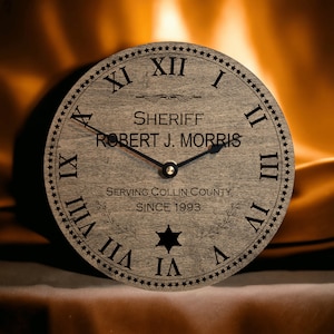 May include: A wooden clock with Roman numerals and a black star in the center. The clock face reads "Sheriff Robert J. Morris Serving Collin County Since 1993".