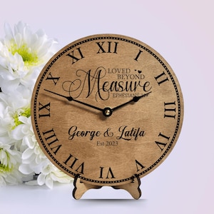 May include: A wooden clock with Roman numeral markings and a black stand. The clock face has the text "Loved Beyond Measure Ephesians 3:19" and "George & Latifa Est 2023".