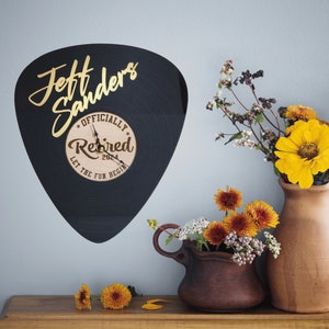 May include: A black guitar pick clock with gold lettering that reads "Jeff Sanders Officially Retired 2024 Let the Fun Begin". The clock is hanging on a light blue wall with a vase of flowers in front of it.