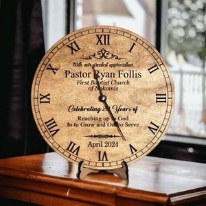 May include: A wooden clock with Roman numerals and a black hand. The clock has a personalized message that reads "With our greatest appreciation Pastor Ryan Follis First Baptist Church of Nokomis Celebrating 20 Years of Reaching up to God In to Grow and Out to Serve April 2024".