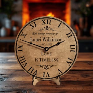 May include: A wooden clock with Roman numerals and a black stand. The clock face reads "In loving memory of Lauri Wilkinson 1967-2023 Love is Timeless".
