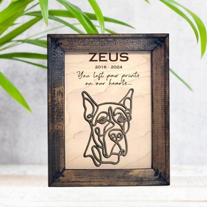 May include: A wooden framed memorial plaque with a brown background and a light brown wood grain pattern. The plaque features a line art illustration of a dog's head and the text "ZEUS 2016-2024 You left paw prints on our hearts..."