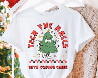 May include: White t-shirt with the text "TECH THE HALLS WITH CODING CHEER" and a cartoon Christmas tree graphic. A red Santa hat hangs nearby. A Christmas tree with lights is in the background. A beige sweater and white pants are also visible.