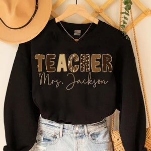 May include: Black sweatshirt with the word "TEACHER" in a patterned design and the name "Mrs. Jackson" in script. The sweatshirt is paired with denim shorts and a tan hat. A woven bag hangs nearby.