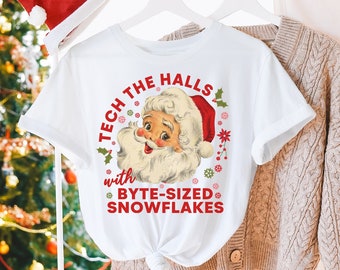 May include: White t-shirt with a Santa Claus graphic and the text "TECH THE HALLS with BYTE-SIZED SNOWFLAKES." A red Santa hat hangs nearby. A Christmas tree with lights is in the background.