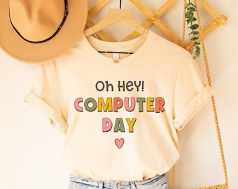 May include: A beige t-shirt with the text "Oh Hey! COMPUTER DAY" in colorful letters and a small pink heart. The shirt is hanging on a wooden hanger with a straw hat and a woven bag.
