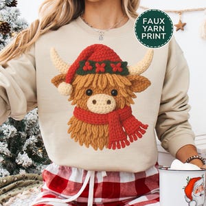 May include: A sand-colored sweatshirt featuring a Highland cow design wearing a red Christmas hat and scarf. The cow has a crocheted appearance. The sweatshirt is paired with red and white plaid pajama pants. The text "FAUX YARN PRINT" is visible.