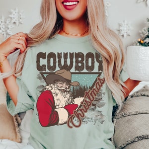 May include: A light blue t-shirt with a vintage-style graphic of Santa Claus wearing a cowboy hat. The graphic says "Cowboy Christmas" in brown text.