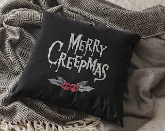 May include: Black square pillow with the text "MERRY CREEPMAS" in a spooky font. Below the text are two red skulls and gray holly leaves. The pillow is on a patterned blanket.
