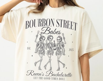 Puede incluir: Camiseta marfil con un gráfico negro que dice "Bourbon Street Babes" con figuras de esqueletos. El texto "Raven's Bachelorette" y "Let the Good Times Roll" también están impresos en la camiseta.