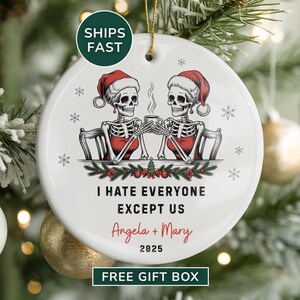 May include: A round white Christmas ornament featuring two skeleton figures wearing Santa hats, toasting with mugs. The ornament has the text "I HATE EVERYONE EXCEPT US" with names and the year 2025. The ornament is decorated with snowflakes and festive greenery.