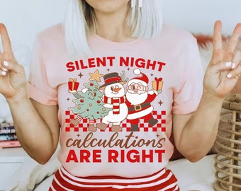 Puede incluir: Camiseta rosa con un diseño navideño con un muñeco de nieve y Papá Noel. El texto de la camiseta dice "Silent Night Calculations Are Right".