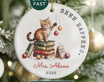 May include: A white ceramic Christmas ornament featuring a tabby cat perched atop a stack of books, reaching for a red ornament. The ornament has the text "SHHH HAPPENS" and "Mrs. Adams 2025".