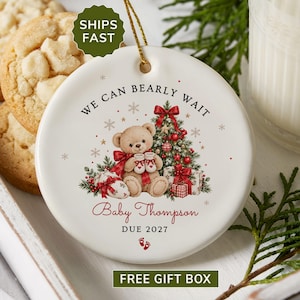 May include: A round white ceramic ornament featuring a teddy bear holding baby booties in front of a Christmas tree. The text reads WE CAN BEARLY WAIT, Baby Thompson, and DUE 2027, with small red baby footprints at the bottom and a gold hanging string.