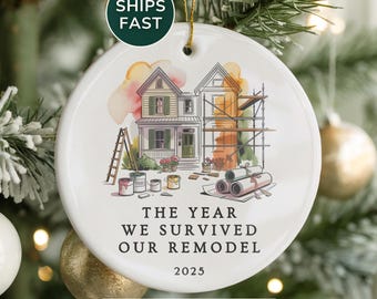 May include: White ceramic Christmas ornament with a watercolor illustration of a house undergoing a remodel. The text reads "THE YEAR WE SURVIVED OUR REMODEL 2025". The ornament hangs from a gold string and is surrounded by Christmas tree branches and ornaments.