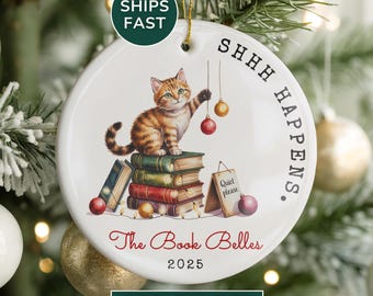 May include: A round ceramic Christmas ornament featuring a tabby cat sitting on books, reaching for ornaments. The ornament has the text "SHHH HAPPENS." and "The Book Belles 2025."