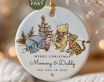 May include: A round white ceramic Christmas ornament featuring a gold hanging cord. The illustration depicts Eeyore, Piglet, Winnie the Pooh, and Tigger near a Christmas tree. Text reads: MERRY CHRISTMAS Mommy & Daddy SEE YOU IN 2027 with two small footprints.