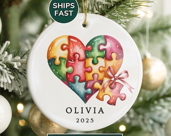 Può includere: Ornamento natalizio in ceramica bianca con un design a cuore a puzzle colorato. L'ornamento ha il nome "OLIVIA" e l'anno "2025" stampati sopra. È appeso a un albero di Natale con ornamenti bianchi e dorati.