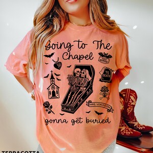 May include: Terracotta-colored t-shirt with a graphic design featuring skeletons in a coffin, bats, and the text "Going to The Chapel gonna get buried". The design includes a church, roses, and a heart pendant.