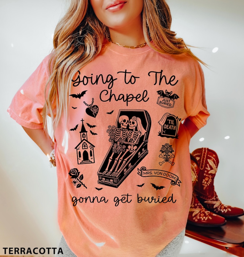 May include: Terracotta-colored t-shirt with a graphic design featuring skeletons in a coffin, bats, and the text "Going to The Chapel gonna get buried". The design includes a church, roses, and a heart pendant.
