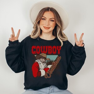 May include: A black sweatshirt with a graphic of a cowboy Santa Claus wearing a red suit and a cowboy hat. The text "COWBOY CHRISTMAS" is printed in red and brown letters.