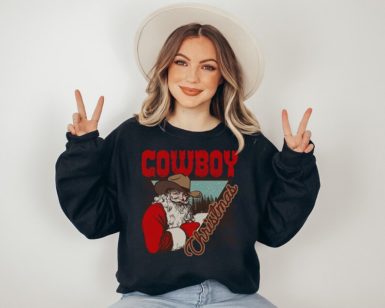 May include: A black sweatshirt with a graphic of a cowboy Santa Claus wearing a red suit and a cowboy hat. The text "COWBOY CHRISTMAS" is printed in red and brown letters.