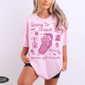 May include: Light pink t-shirt with a graphic design in a darker pink color. The design includes the text "Going to the Chapel gonna get buried" with illustrations of bats, a church, a coffin with skeletons, and other gothic elements.