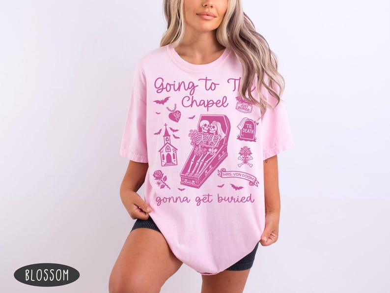 May include: Light pink t-shirt with a graphic design in a darker pink color. The design includes the text "Going to the Chapel gonna get buried" with illustrations of bats, a church, a coffin with skeletons, and other gothic elements.