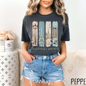 May include: A dark gray t-shirt with a graphic print of a Parisian cityscape, including the Eiffel Tower, a bicycle, and streetlights. The text "Paris is always a good idea!" is printed below the graphic.