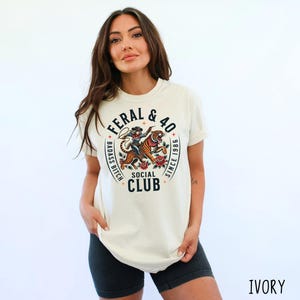 May include: Ivory-colored t-shirt with a circular graphic featuring a tiger and a figure. The text reads "FERAL & 40 BADASS BITCH SOCIAL CLUB SINCE 1986". The shirt is worn with black shorts.